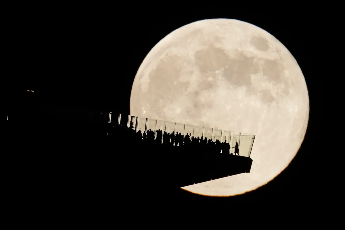 A large, detailed full moon dominates the night sky, with the silhouettes of many people standing on a modern glass-railed…