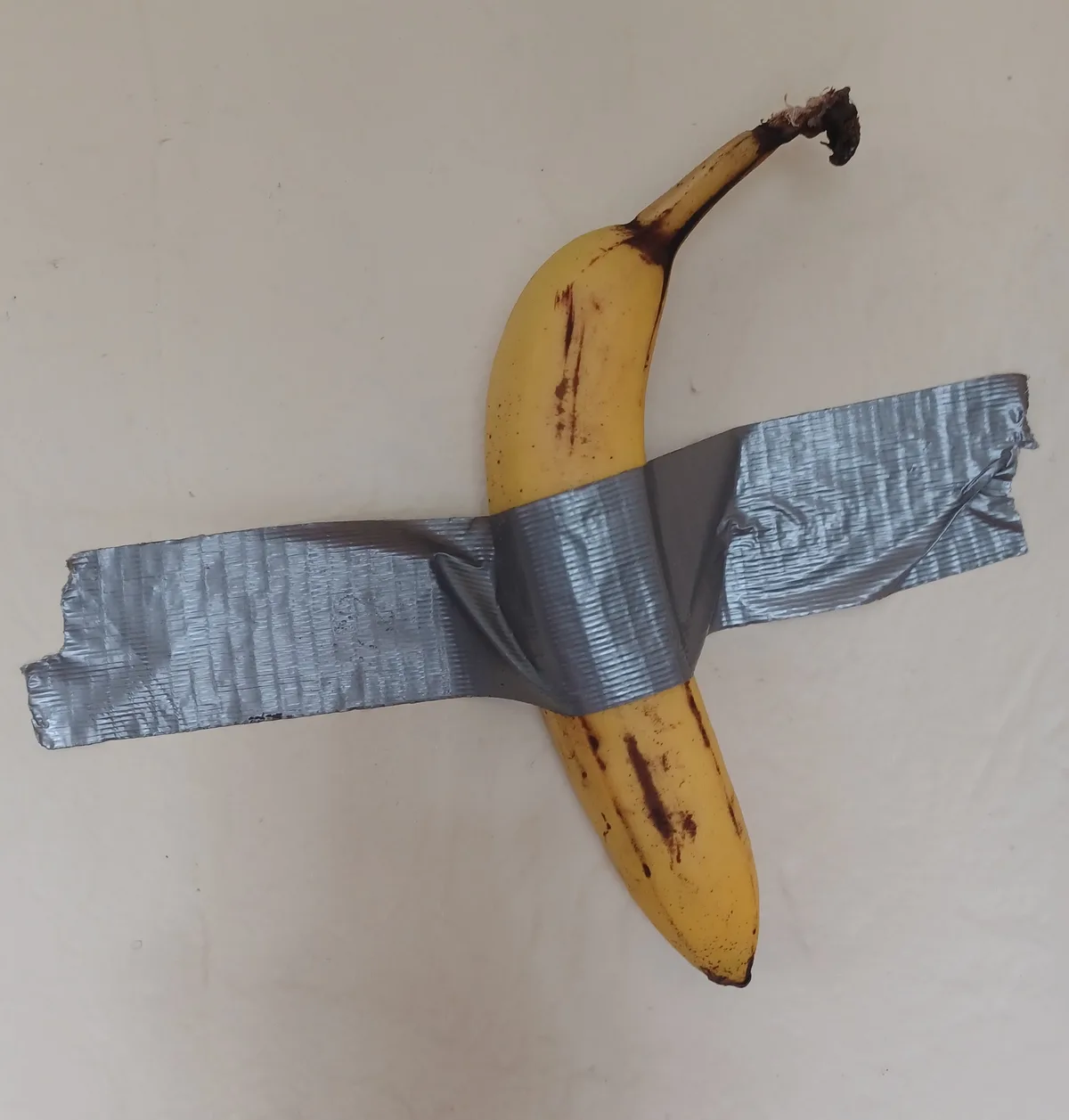 A ripe yellow banana with brown spots lies on a plain off-white surface, its stem pointing upward. A wide strip of silver…