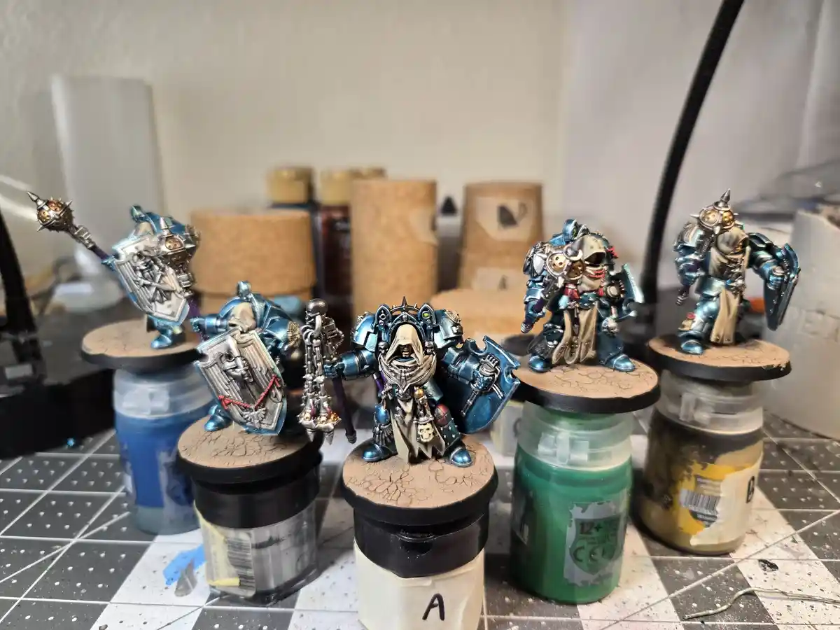 Five intricately painted miniature fantasy warriors in blue and silver armor stand on bases atop paint jars on a checkered…