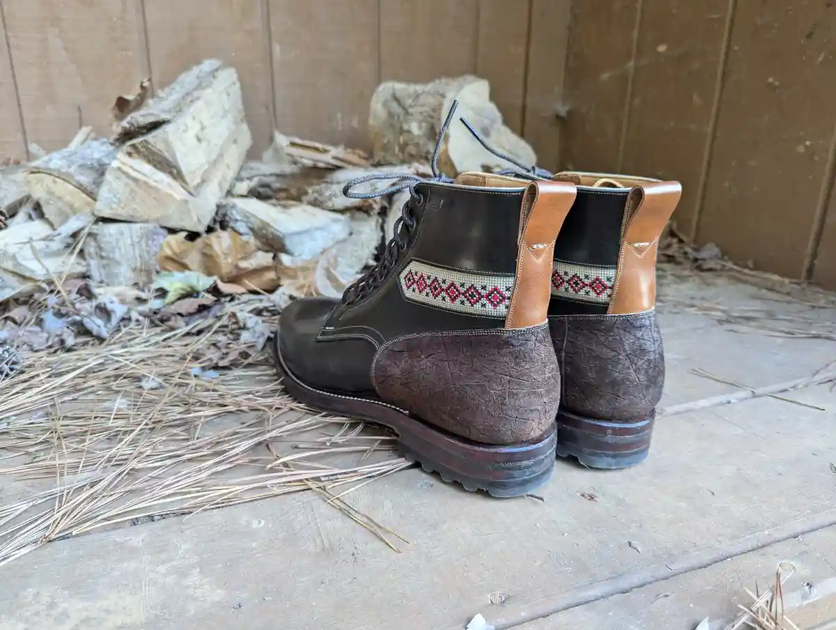A pair of rugged brown leather boots with red and black geometric embroidery on the back, standing on a wooden floor covered…