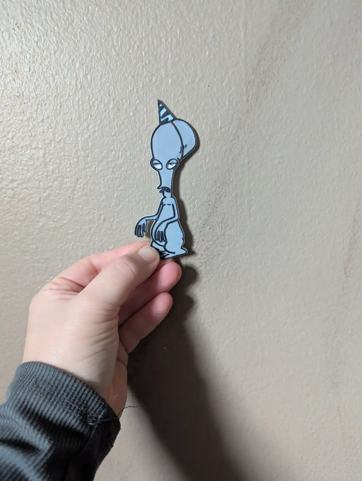 A hand holds a light blue, 3D-printed cutout of the cartoon alien character from 'The Oblongs' against a beige wall. The…