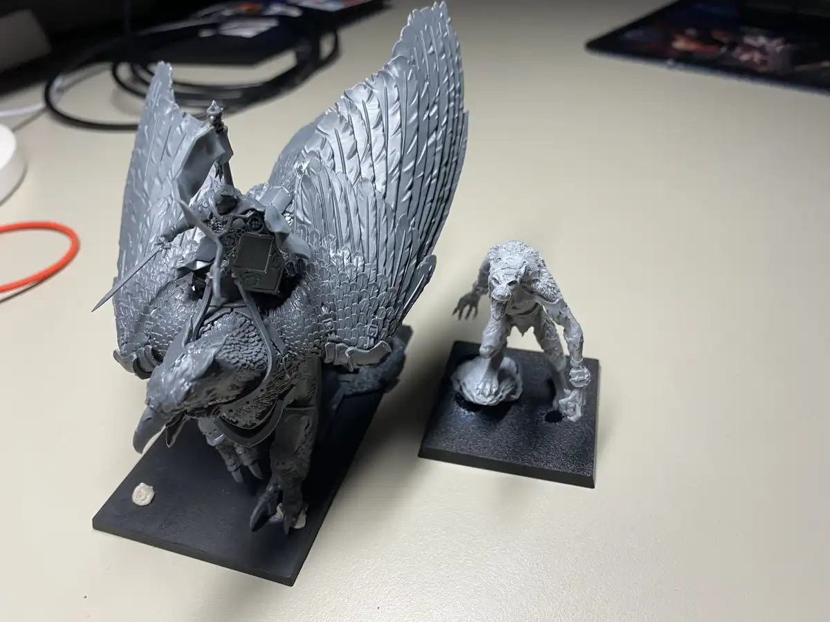 Two unpainted fantasy miniatures sit on a desk: a large griffin with a rider and spread wings, and a smaller, crouching…