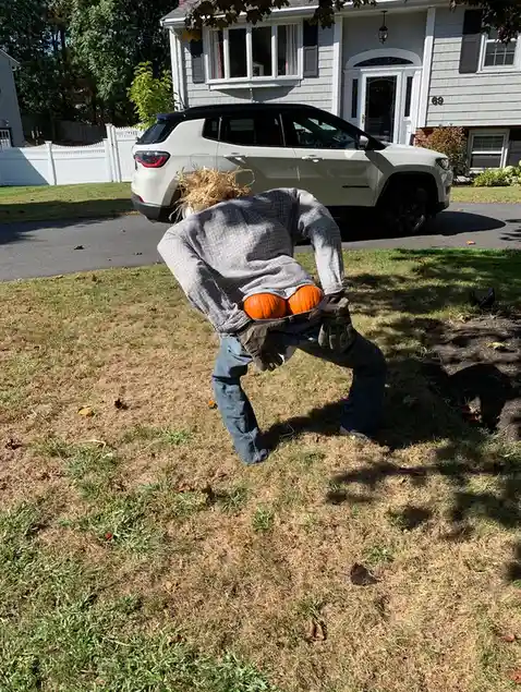 A person in a plaid shirt and jeans is bent over in a grassy yard, holding two small pumpkins in their gloved hands. Behind…