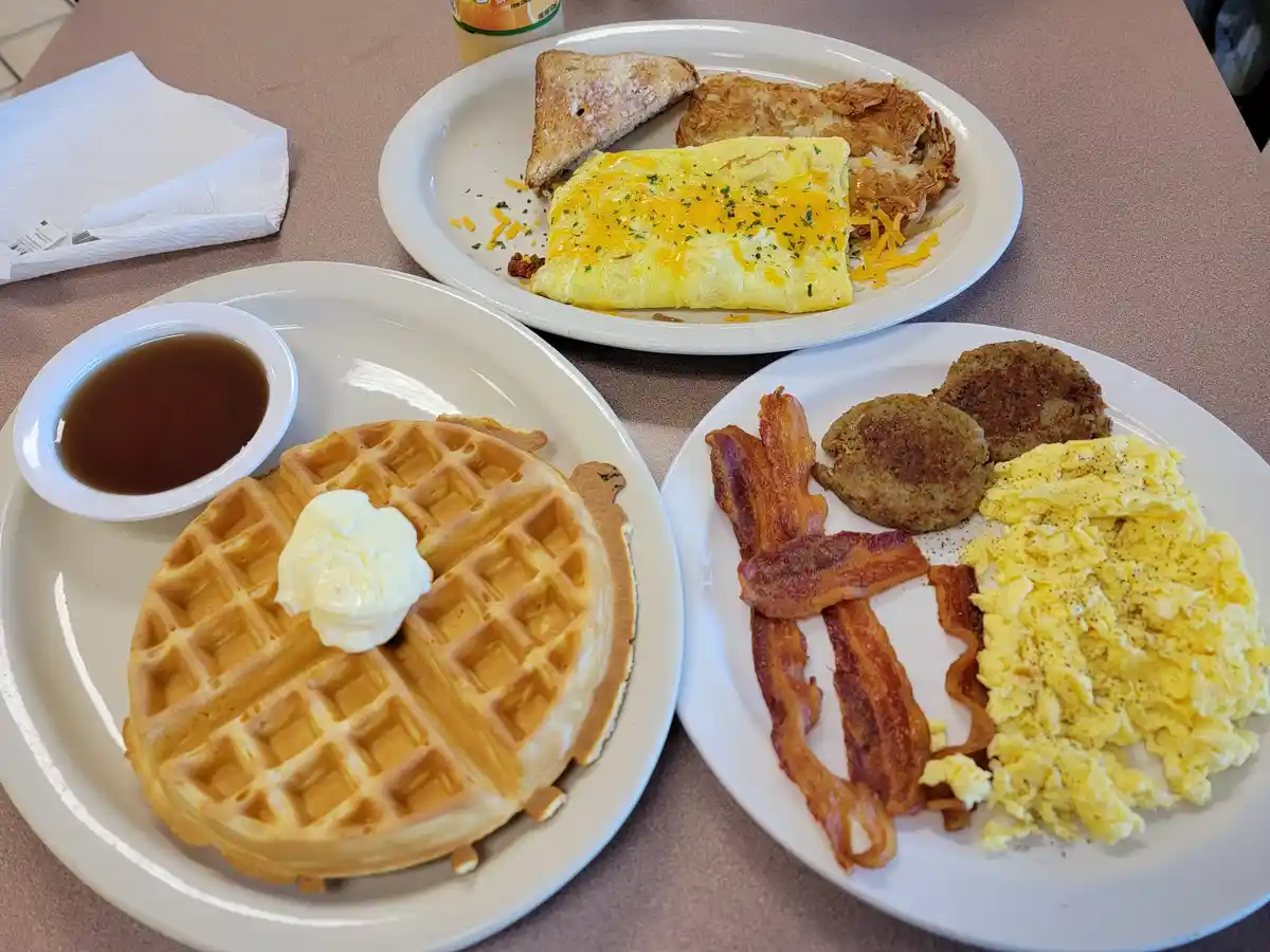 Three plates of breakfast food are arranged on a table: one with a waffle topped with butter and a side of syrup, another…