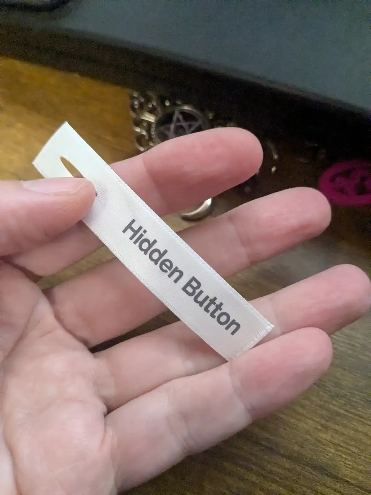 A hand holds a white fabric clothing tag with the text 'Hidden Button' printed on it. The background shows a wooden surface…
