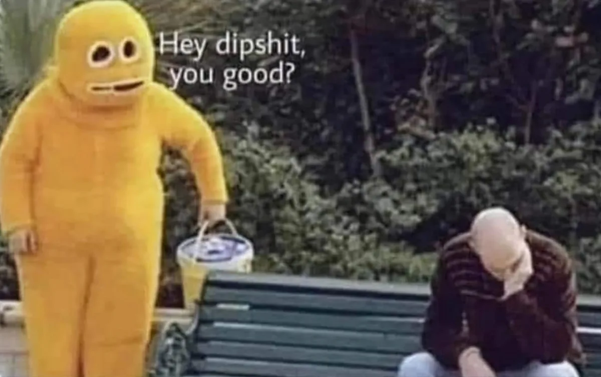 A person in a full-body yellow mascot costume with large, simple eyes and a mouth stands next to a park bench, holding a…