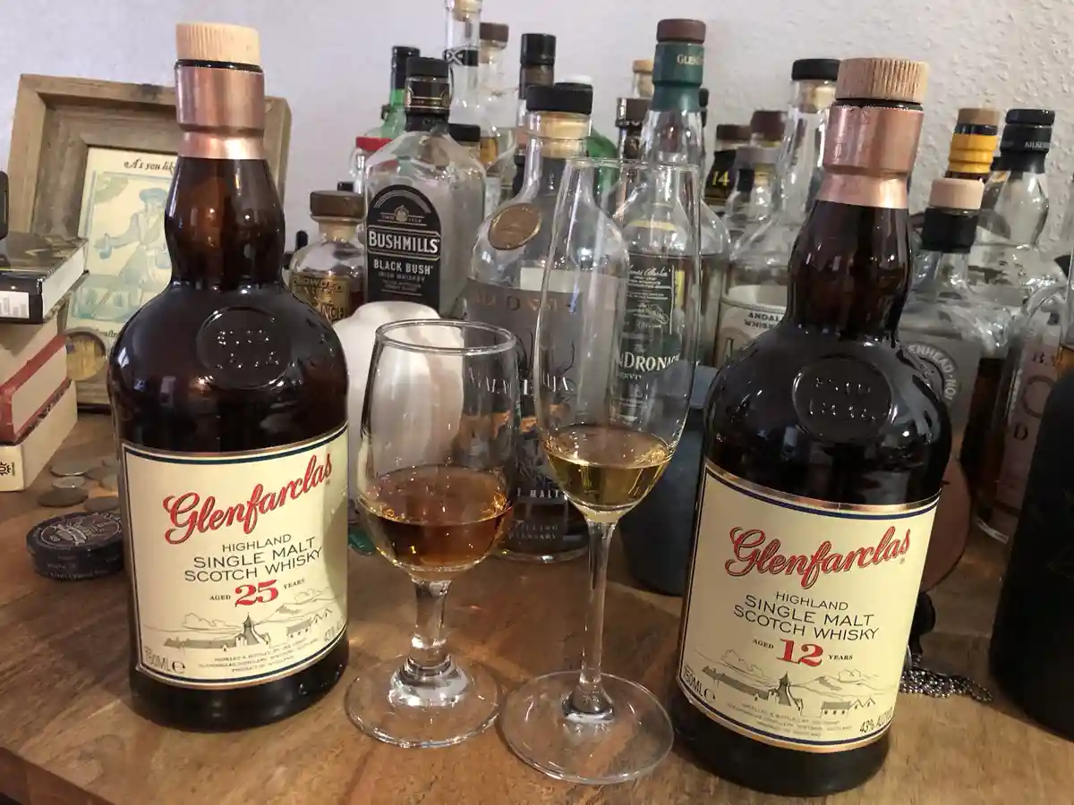 Two Glenfarclas whisky bottles, one aged 25 years and one aged 12 years, sit on a wooden table with two glasses of whisky…