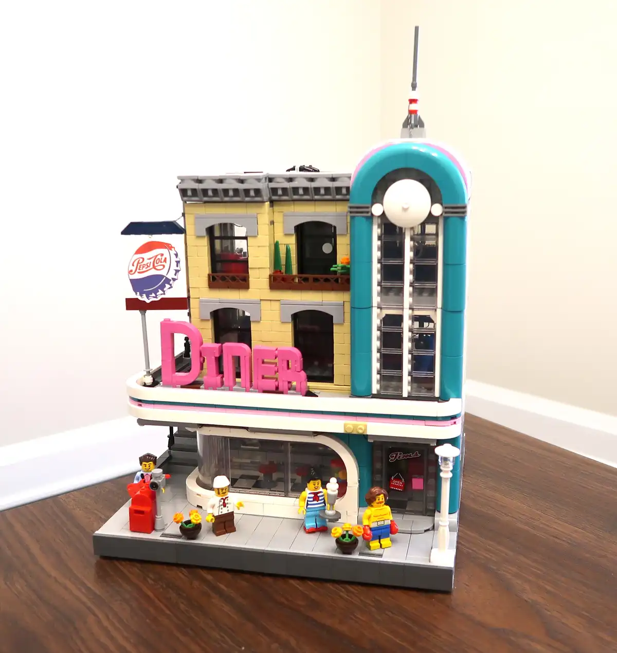 A detailed LEGO model of a retro-style diner with a yellow brick facade, pink 'DINER' sign, and a turquoise tower with a…