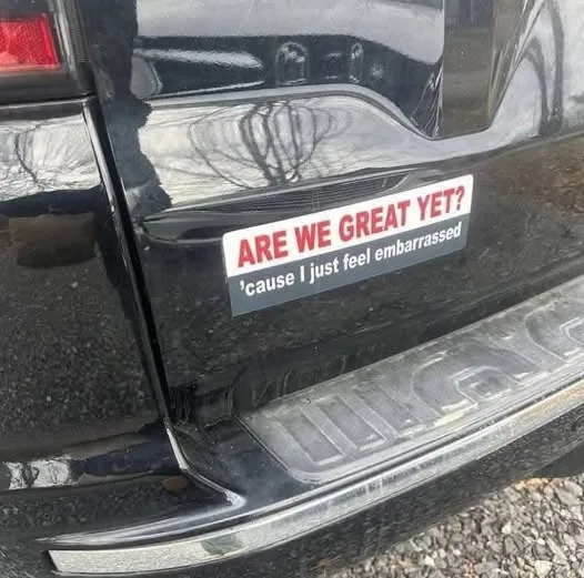 A close-up photo of a black SUV's rear bumper shows a white bumper sticker with red and black text. The top line reads 'ARE…