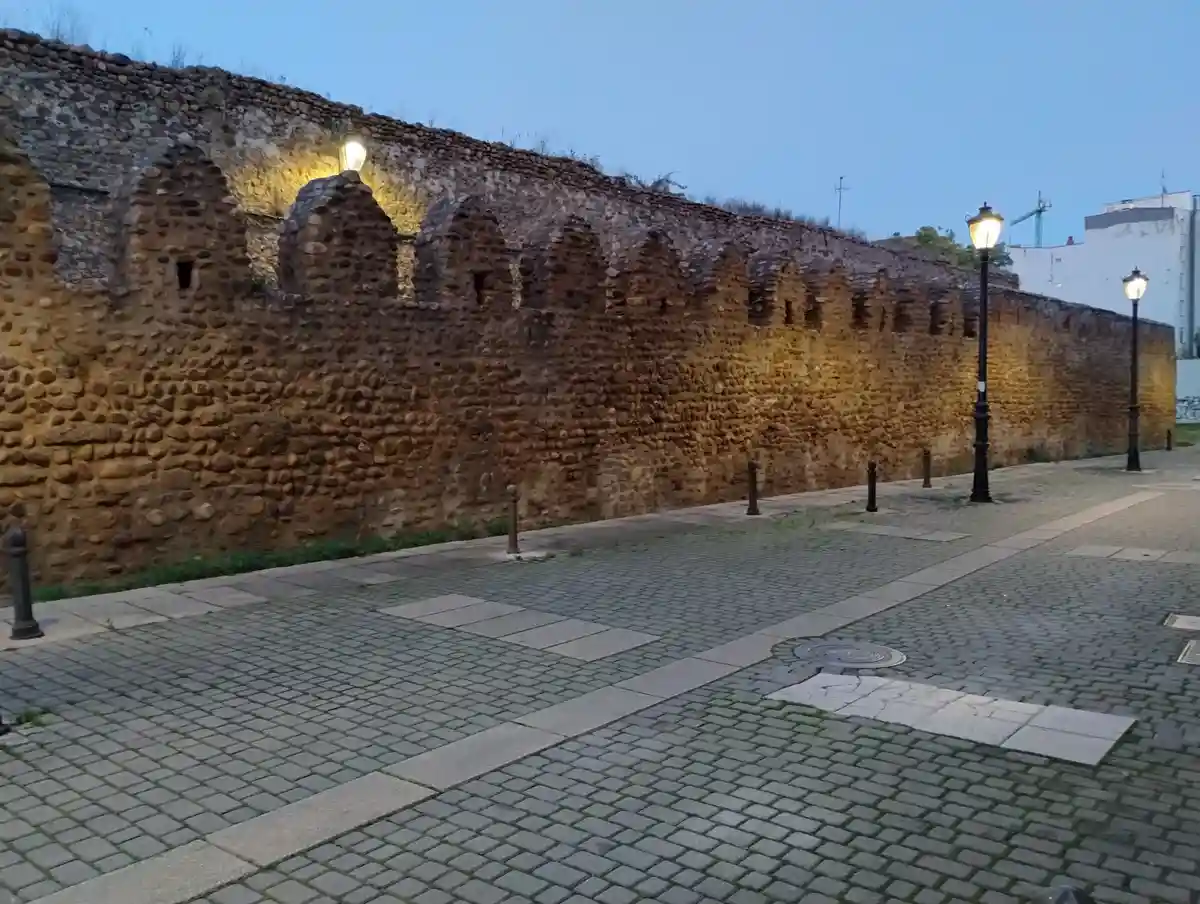 A photo of a long, ancient stone wall with crenellations, illuminated by street lamps at dusk, alongside a paved walkway…