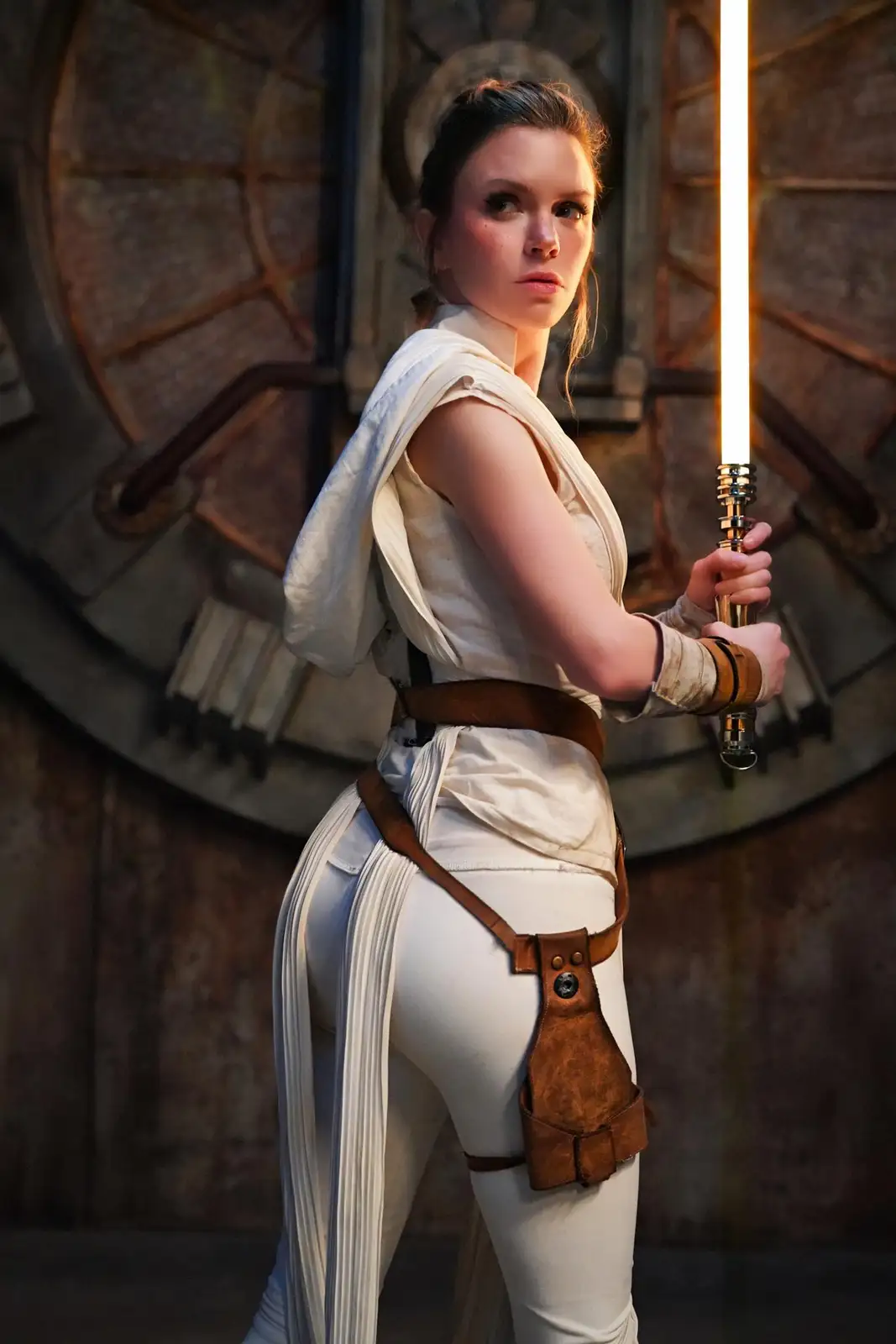 A woman in a white Jedi outfit from Star Wars holds a glowing lightsaber, looking over her shoulder with a serious…