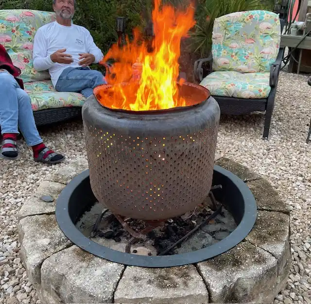 A man sits on a flamingo-patterned outdoor couch beside a large, metal fire pit with bright orange flames erupting from it…