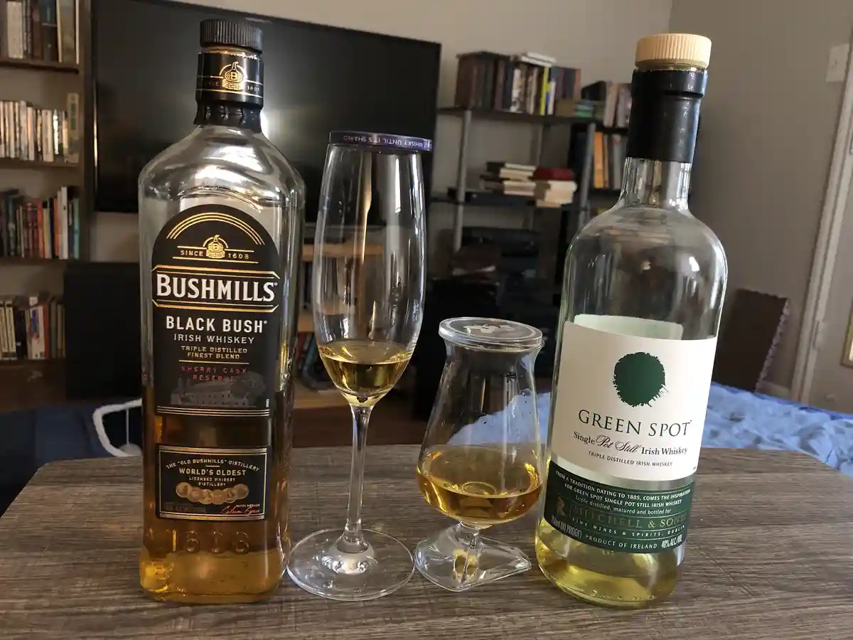 Two bottles of Irish whiskey, Bushmills Black Bush and Green Spot, sit on a wooden table with two glasses filled with…