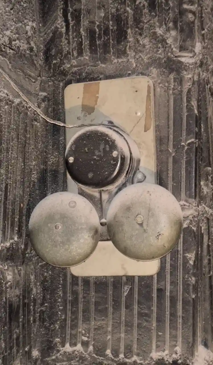 A close-up of a weathered, three-bell doorbell assembly mounted on a textured, snow-dusted wall. The central bell is dark…