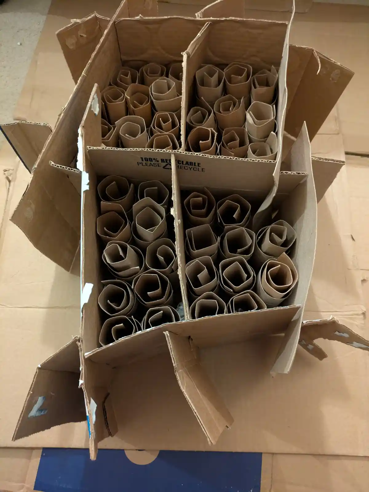 An overhead shot of an open cardboard box divided into four compartments, each filled with rolled-up cardboard tubes…