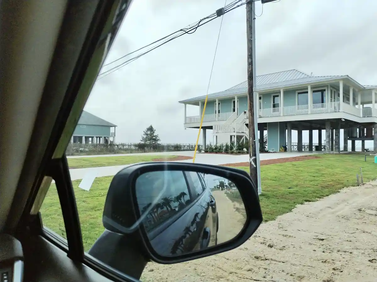A photo taken from inside a car shows a view through the passenger-side window and side mirror. The mirror reflects a sandy…