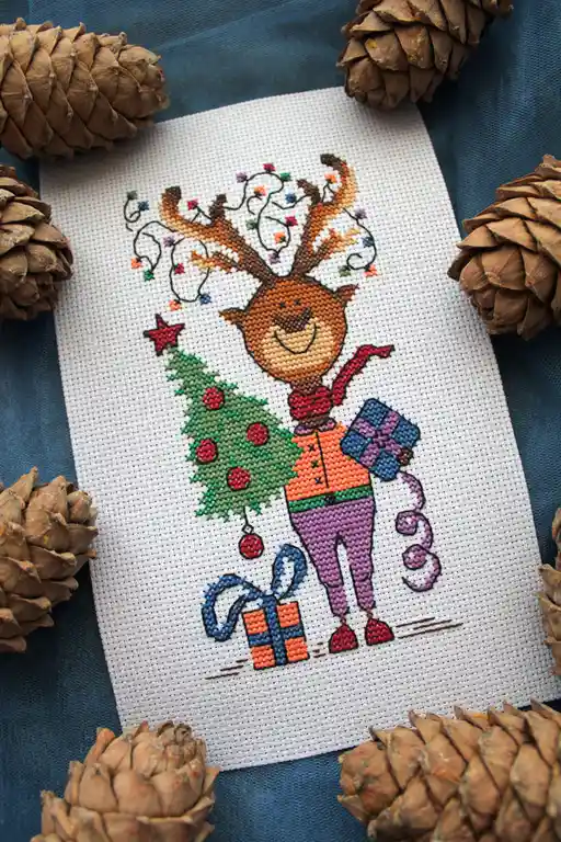A cross-stitch pattern depicts a smiling deer wearing a red scarf and holding a small decorated Christmas tree and a wrapped…