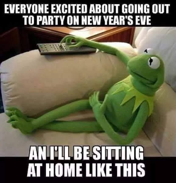 A green Kermit the Frog plush toy lounges lazily on a beige couch, one arm extended to hold a remote control, while the…