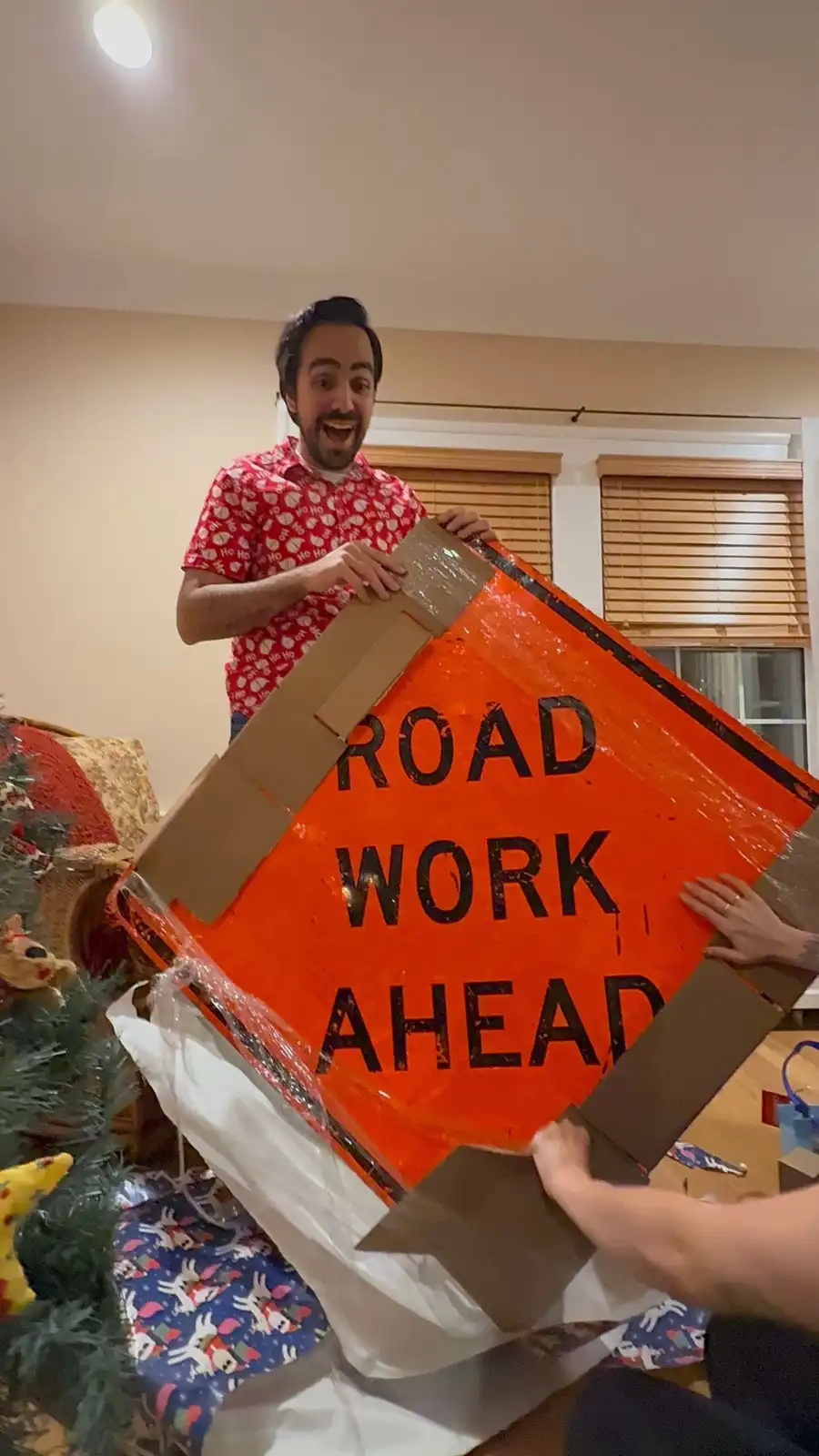 A man in a red 'ho ho ho' patterned shirt stands indoors, grinning widely while holding up a large, orange, diamond-shaped…