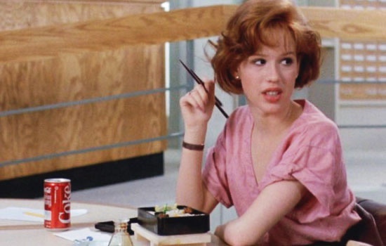 A woman with short red hair sits at a table with a sushi box and a can of Coca-Cola, holding chopsticks up to her face with…