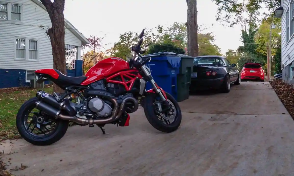 A red Ducati Monster motorcycle is parked on a residential driveway next to a blue trash bin, with a black Mazda MX-5 Miata…