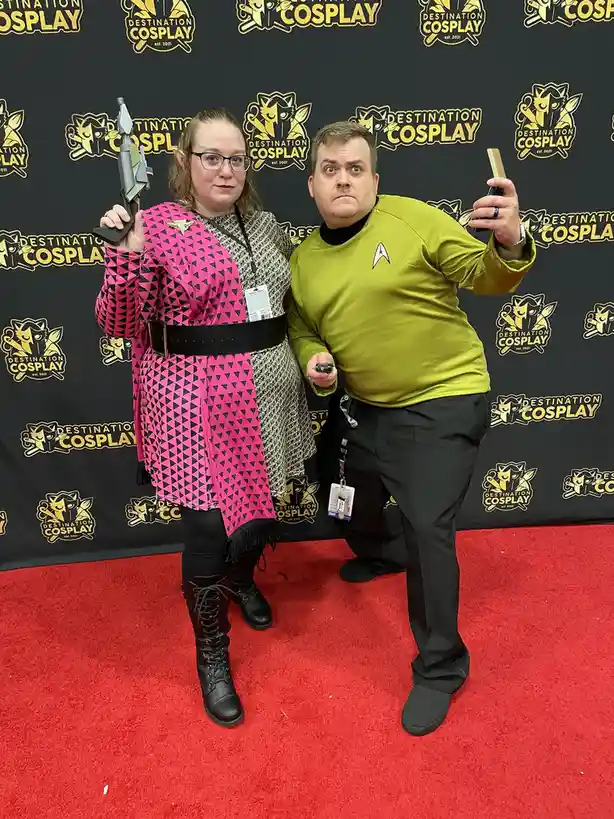 Two people in cosplay stand on a red carpet in front of a 'Destination Cosplay' backdrop. The woman wears a pink and black…