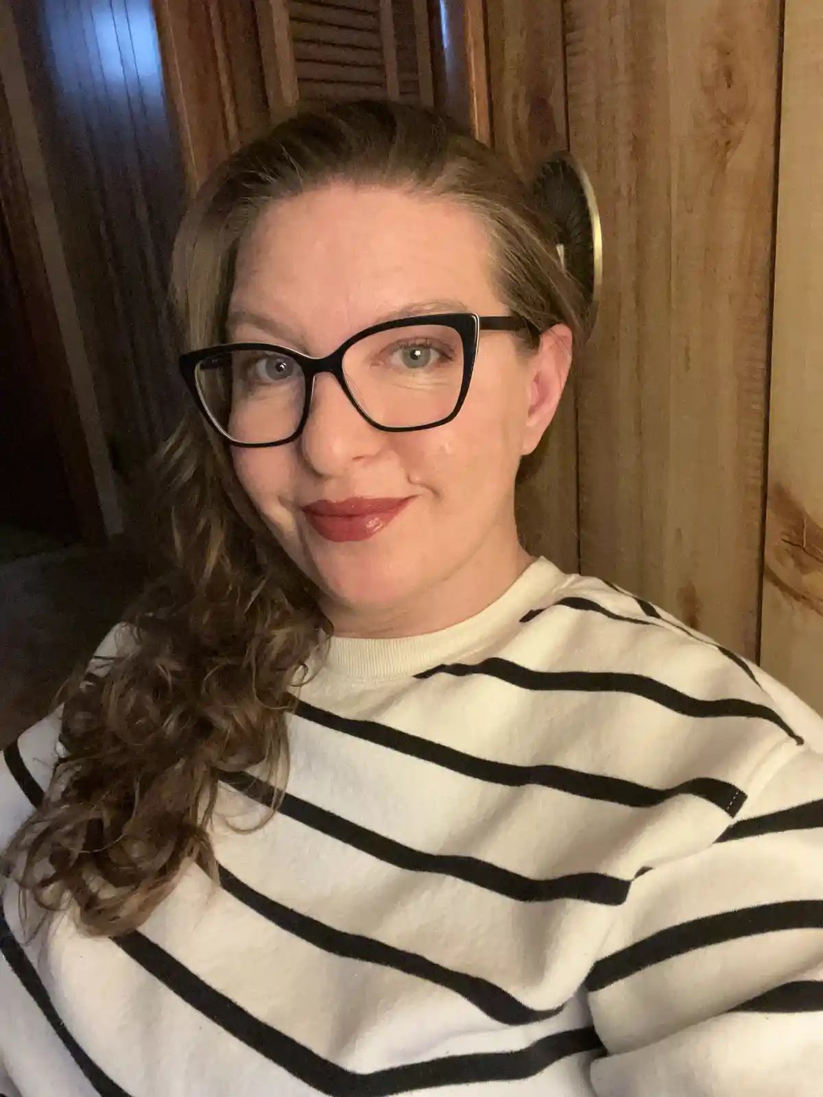 A woman with curly brown hair, wearing black-rimmed glasses and a black-and-white striped sweater, smiles at the camera…