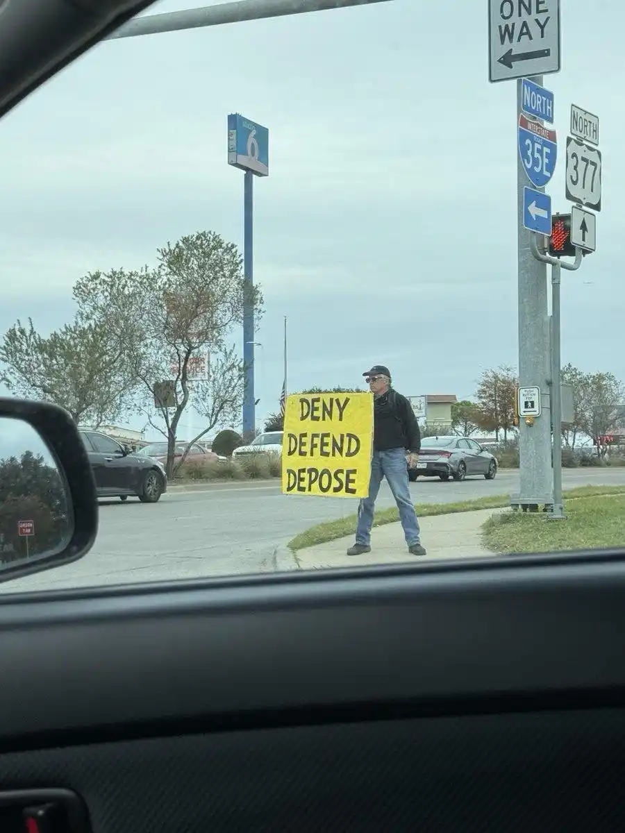 A man stands on a sidewalk holding a yellow sign with the words 'DENY DEFEND DEPOSE' in bold black letters. He is wearing a…