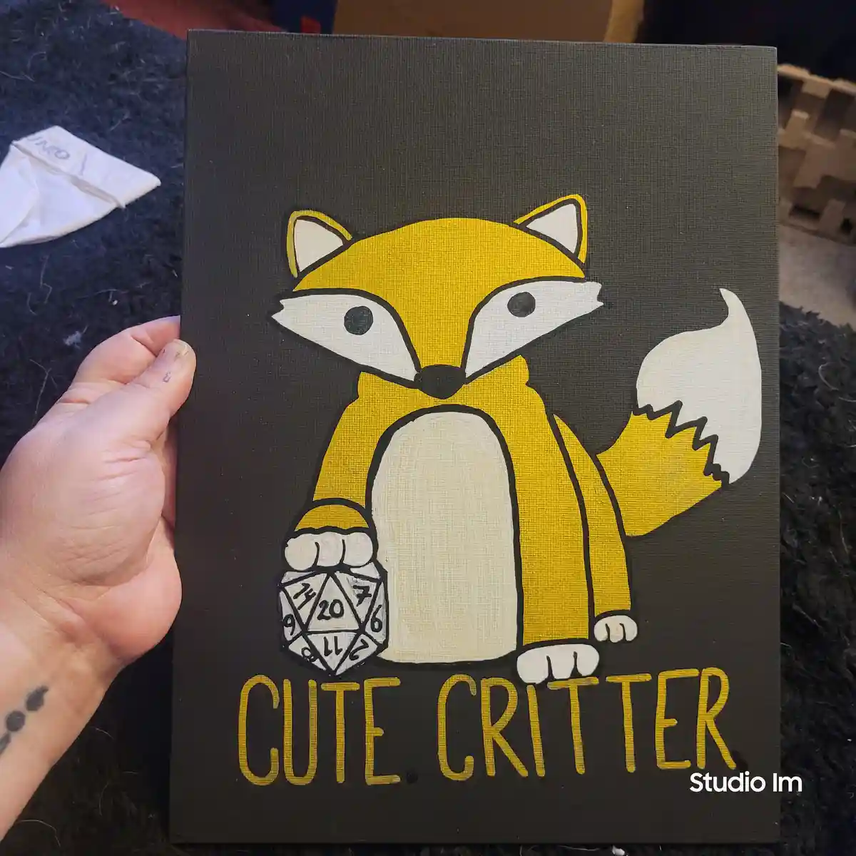 A hand holds a dark gray canvas painting featuring a cartoon-style yellow fox with a white-tipped tail and chest, holding a…