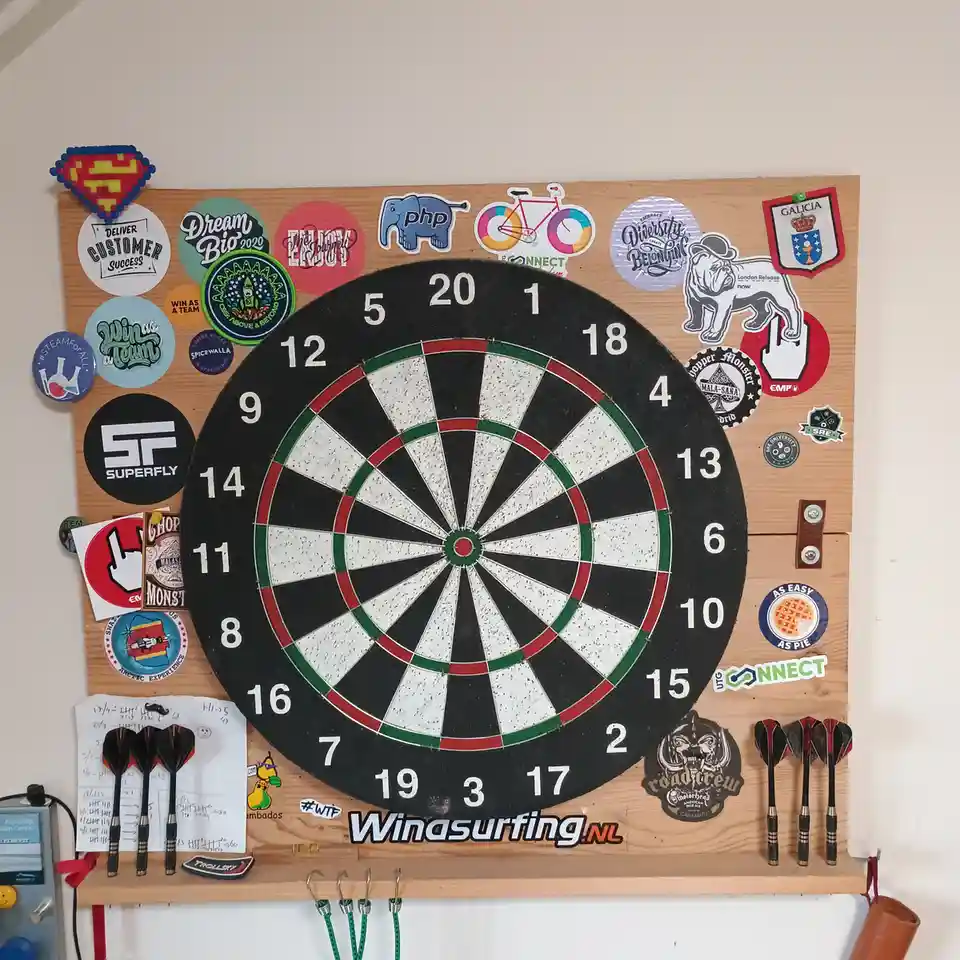 A dartboard mounted on a wooden board covered in numerous colorful stickers, with darts resting on hooks below. The board…