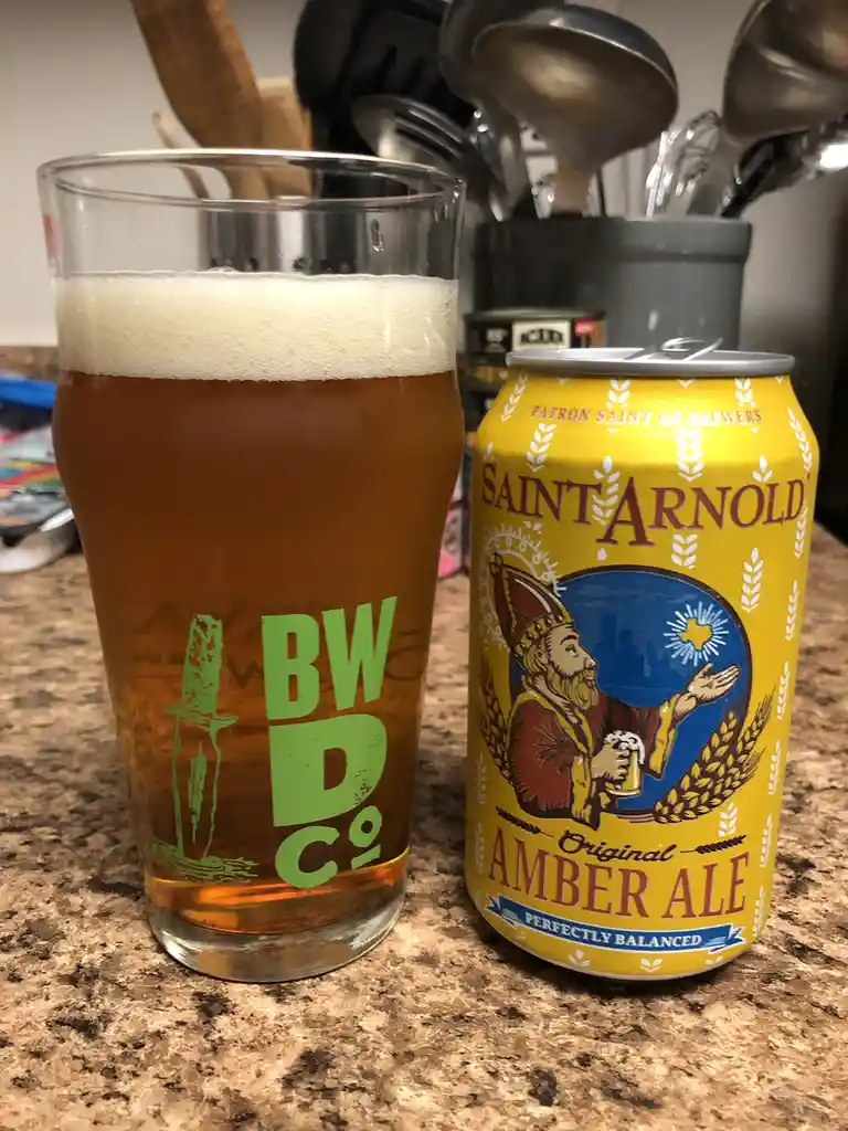 A glass of amber ale with a white head sits next to a yellow can of Saint Arnold Original Amber Ale on a speckled…
