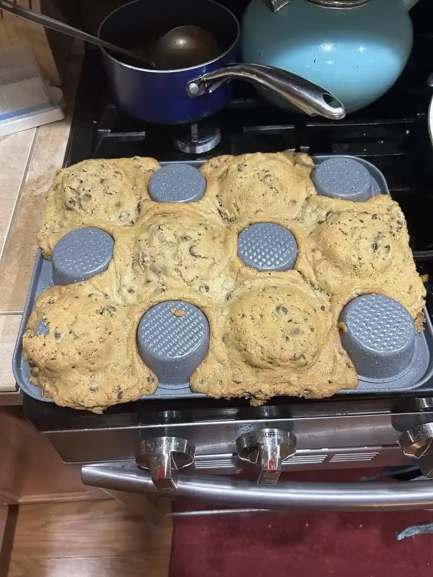 A close-up photo shows a muffin tin on a stovetop with partially baked chocolate chip cookies that have overflowed and…