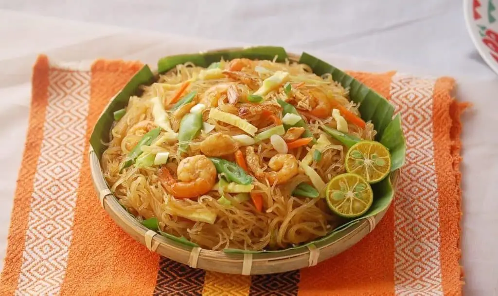 A close-up photo of a bowl of stir-fried rice noodles with shrimp, vegetables, and lime wedges, served on a banana leaf in a…