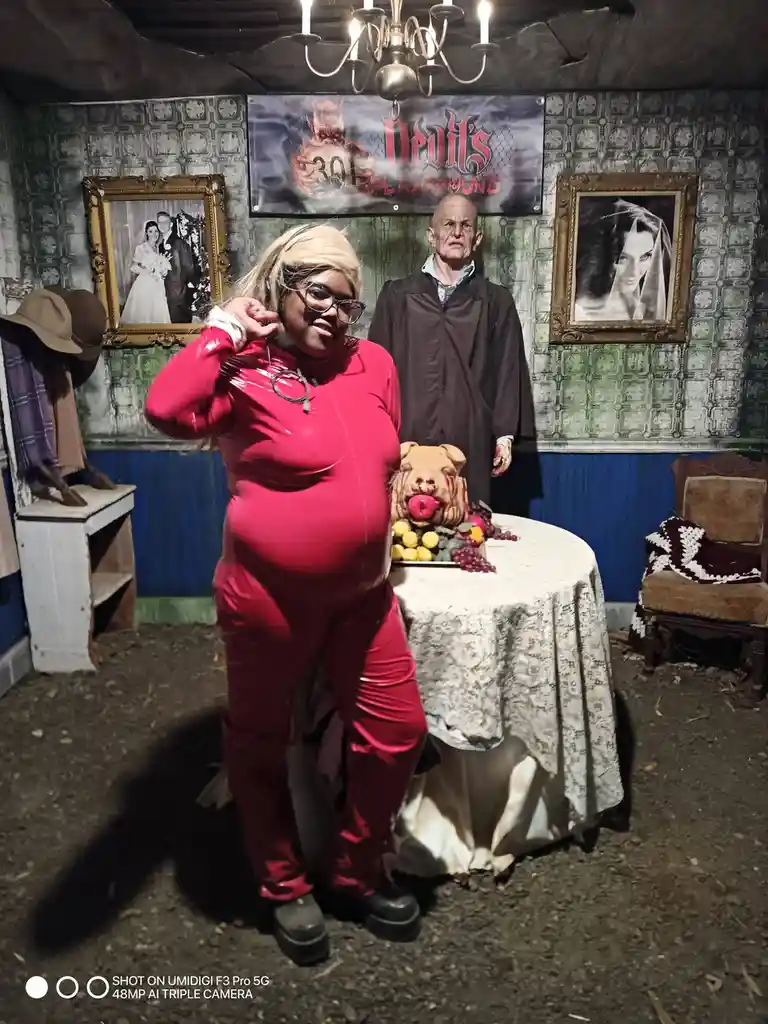 A person in a shiny red jumpsuit and glasses poses confidently in front of a horror-themed set labeled 'Devil's Playground,'…