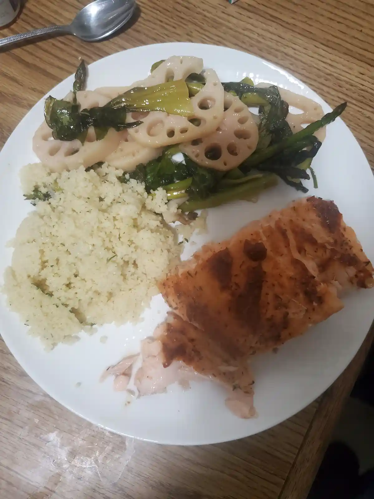 A white plate on a wooden table holds a meal of grilled salmon, couscous, and stir-fried lotus root and greens. The salmon…
