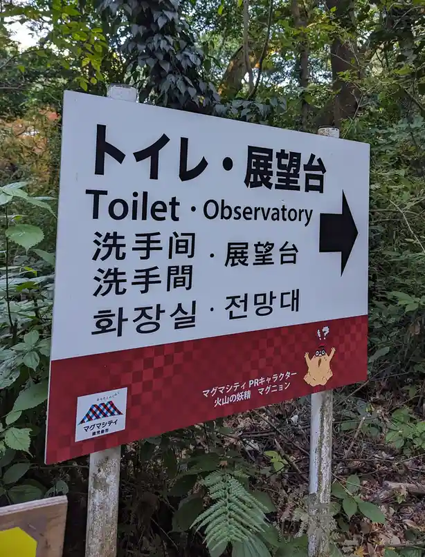 A white directional sign in a forest points right with an arrow, indicating 'Toilet · Observatory' in Japanese, Chinese, and…