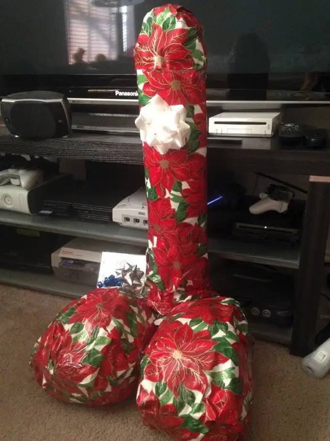 a large, phallic-shaped object is wrapped in red and green poinsettia-patterned wrapping paper with a white bow at the top…