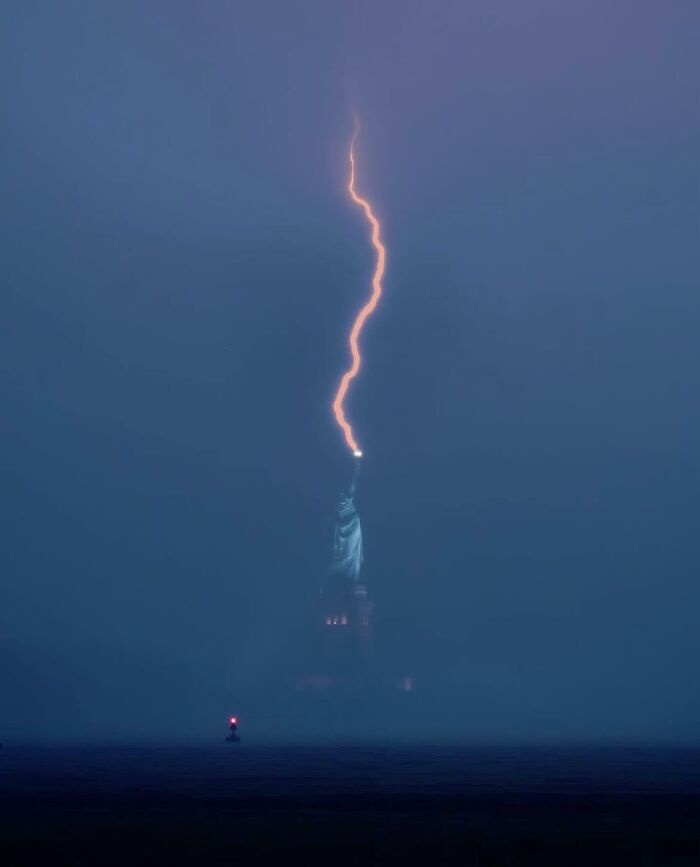 A dark, foggy image shows the Statue of Liberty in the distance, with a bright, jagged lightning bolt striking down from the…