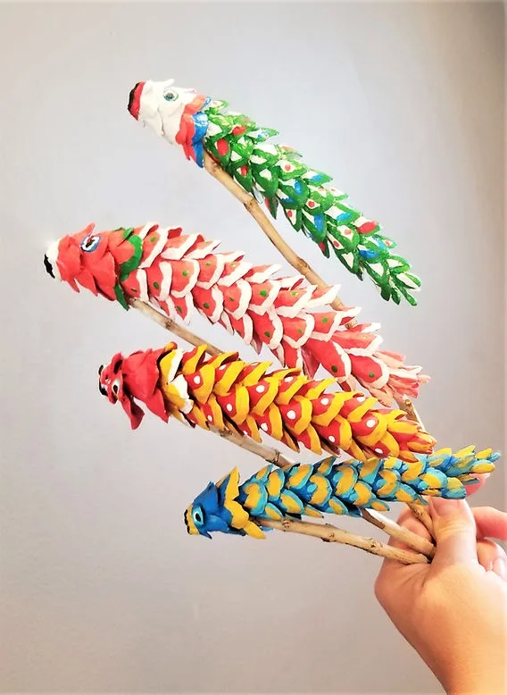 A hand holds four colorful, handcrafted fish-shaped decorations made from painted pinecones, each mounted on a twig and…