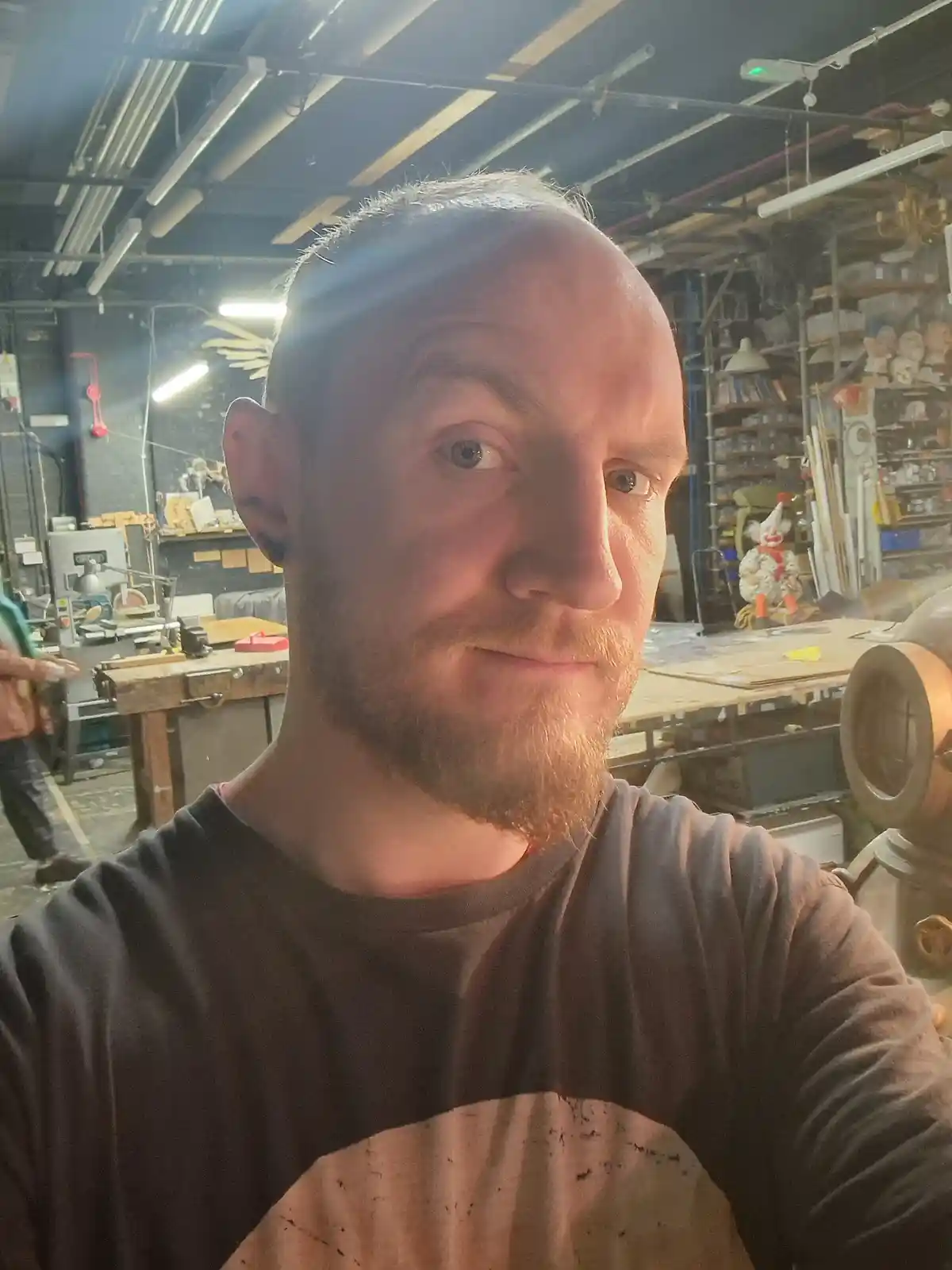 A man with a beard and short hair takes a selfie in a cluttered workshop filled with tools, shelves of materials, and a…