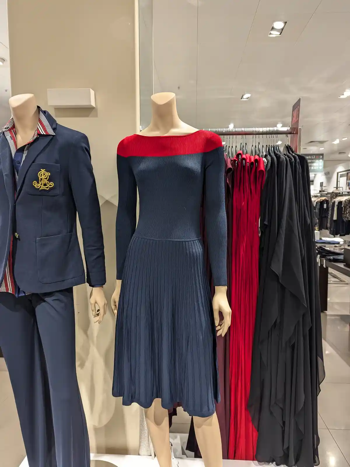 Two mannequins stand in a clothing store; one wears a blue suit with a striped shirt and a gold embroidered logo, the other…