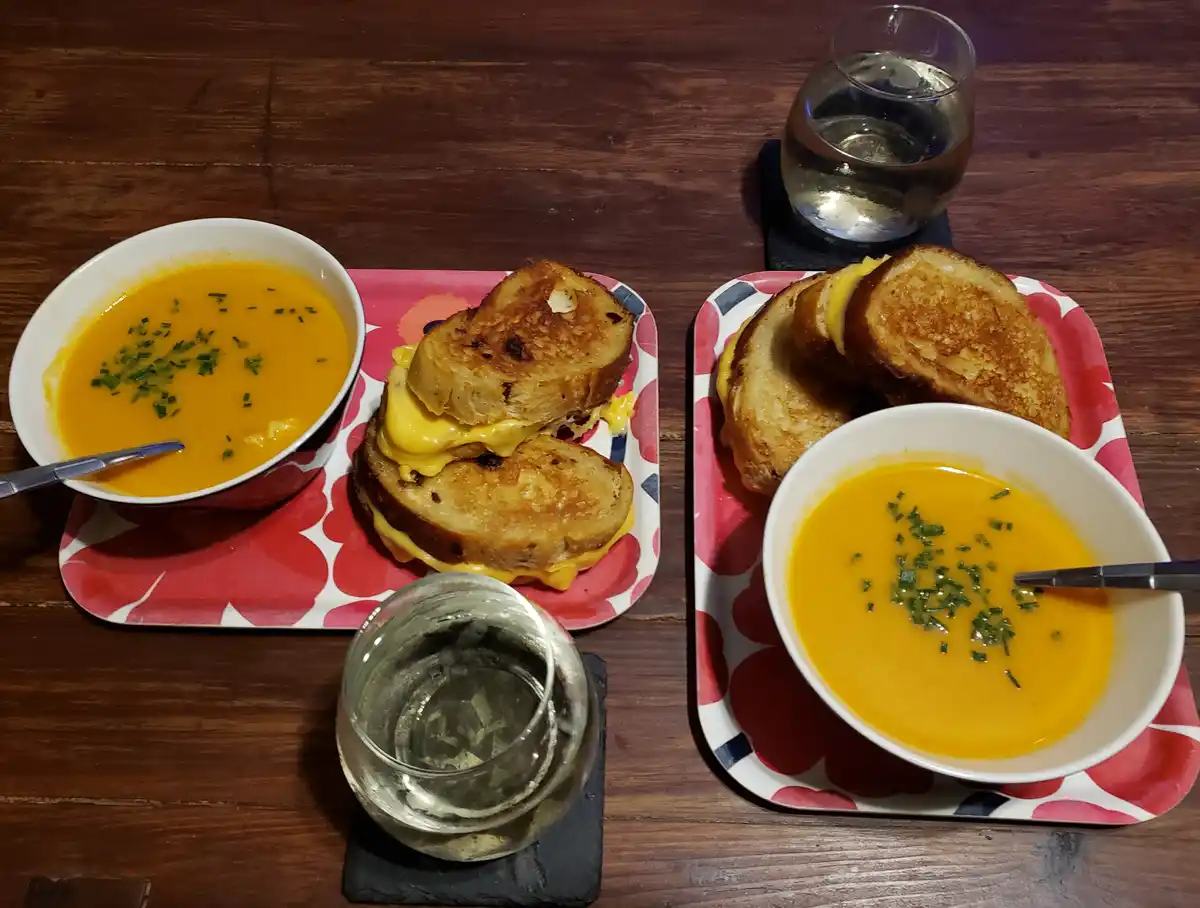 Two identical meals, each consisting of a bowl of orange soup garnished with chopped green herbs and a grilled cheese…