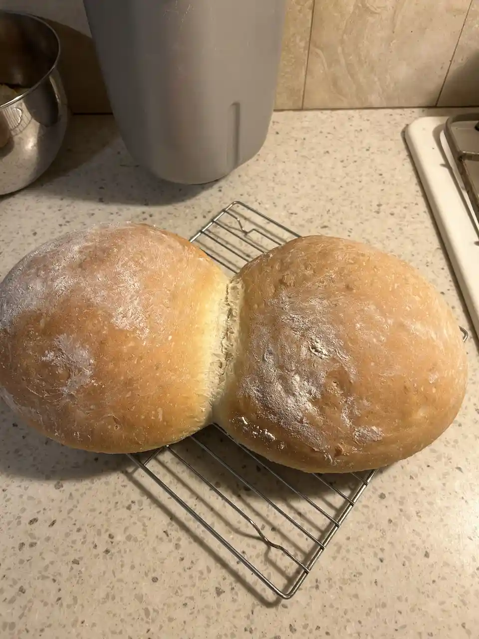 Two freshly baked, golden-brown bread rolls sit side-by-side on a metal cooling rack on a speckled kitchen countertop…