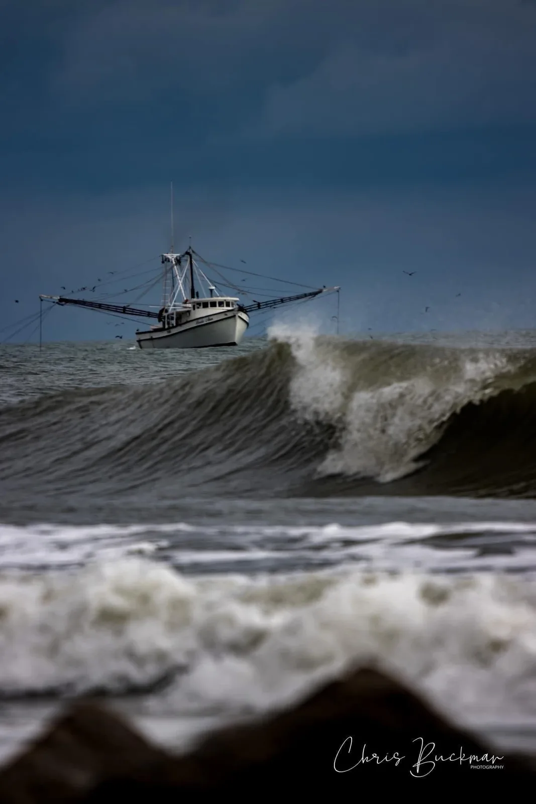 A white shrimp boat sails calmly in the distance under a dark, stormy sky, while a large, crashing wave dominates the…