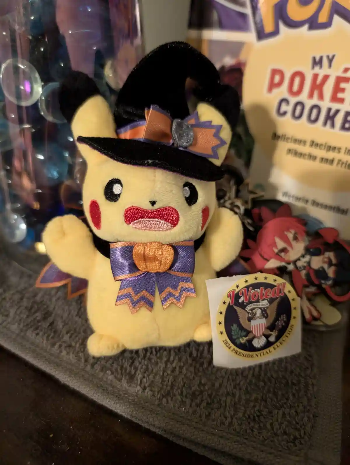 A plush Pikachu dressed in a Halloween witch costume with a black hat and purple-orange ribbon, standing next to a 2024…