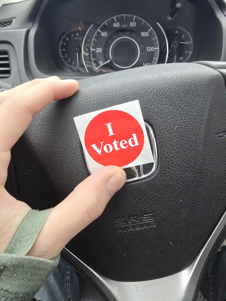 A person's hand holds a red circular 'I Voted' sticker over the airbag cover of a car's steering wheel, with the dashboard…