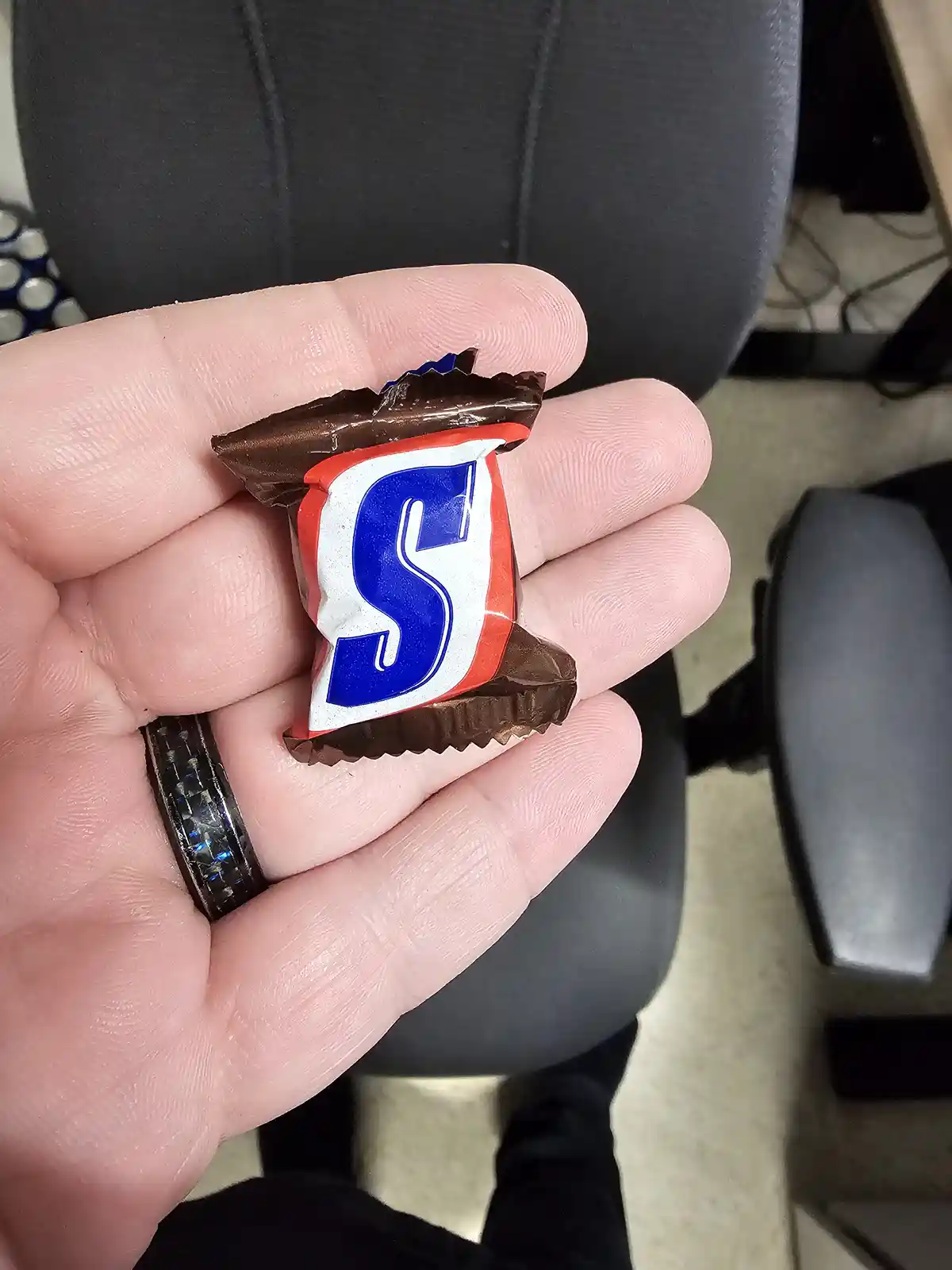 A hand holds a small, partially unwrapped chocolate candy with a wrapper featuring a large blue 'S' on a white and red…