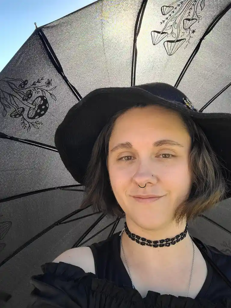 A person with short dark hair, a black hat, and a nose ring smiles at the camera while holding a large, open, silver-colored…