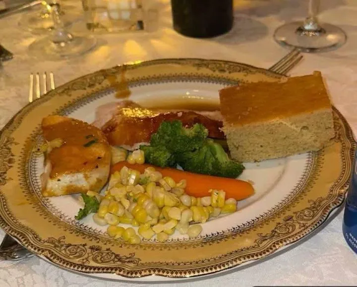 A plated meal featuring chicken, broccoli, corn, a carrot, and a slice of cornbread on an ornate gold-rimmed plate. The…