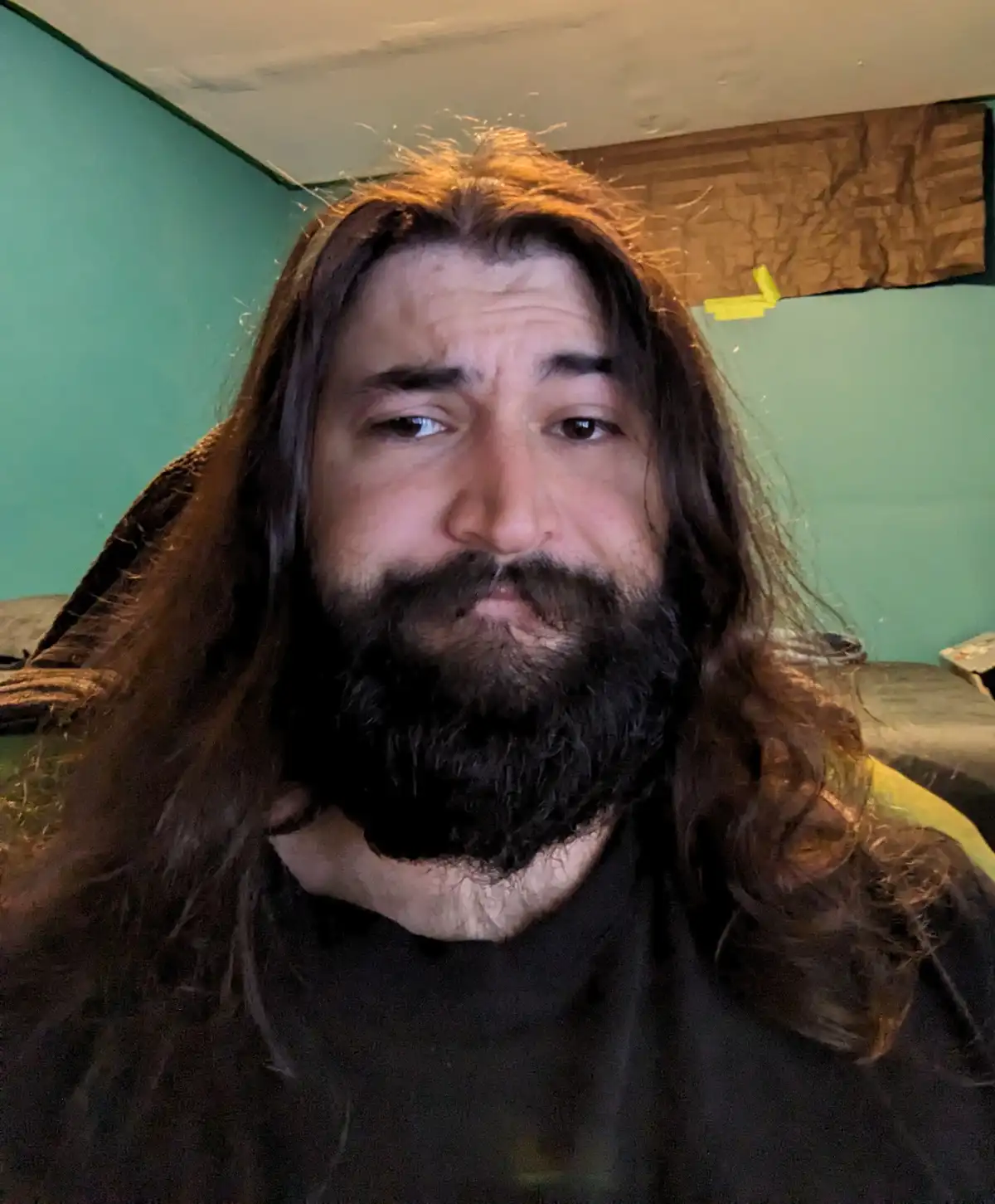 A man with long brown hair and a full beard stares directly at the camera with a slightly furrowed brow and a neutral…